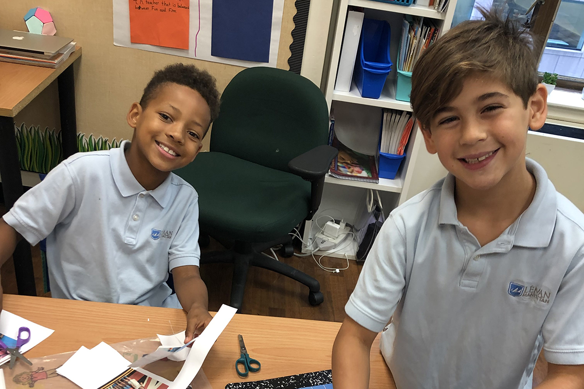 Two boys sit smile as they sit at desks in their classroom