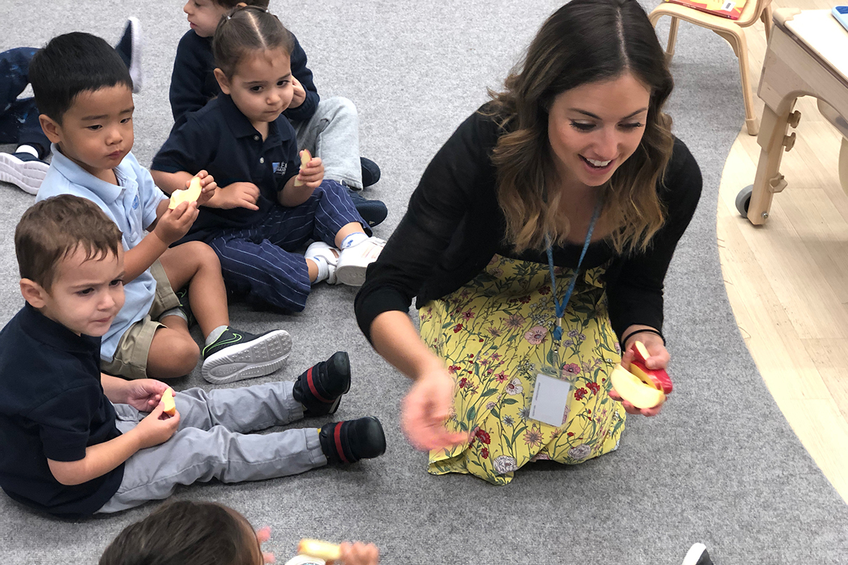 A teacher and students sit on the floor eating apples